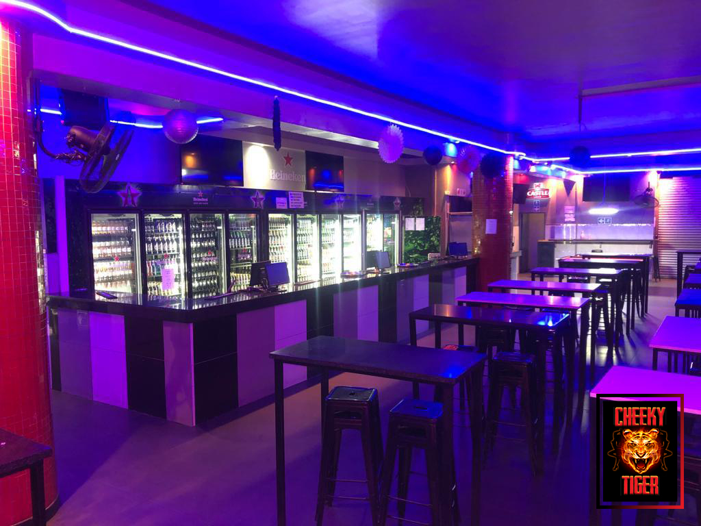 Main bar with stocked fridges and high tables under purple neon at Cheeky Tiger Noord Street